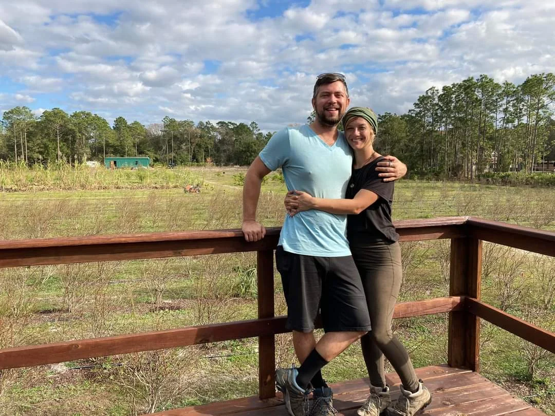Laura and Steve on the orchard deck with the blueberry rows and tall pines behind them